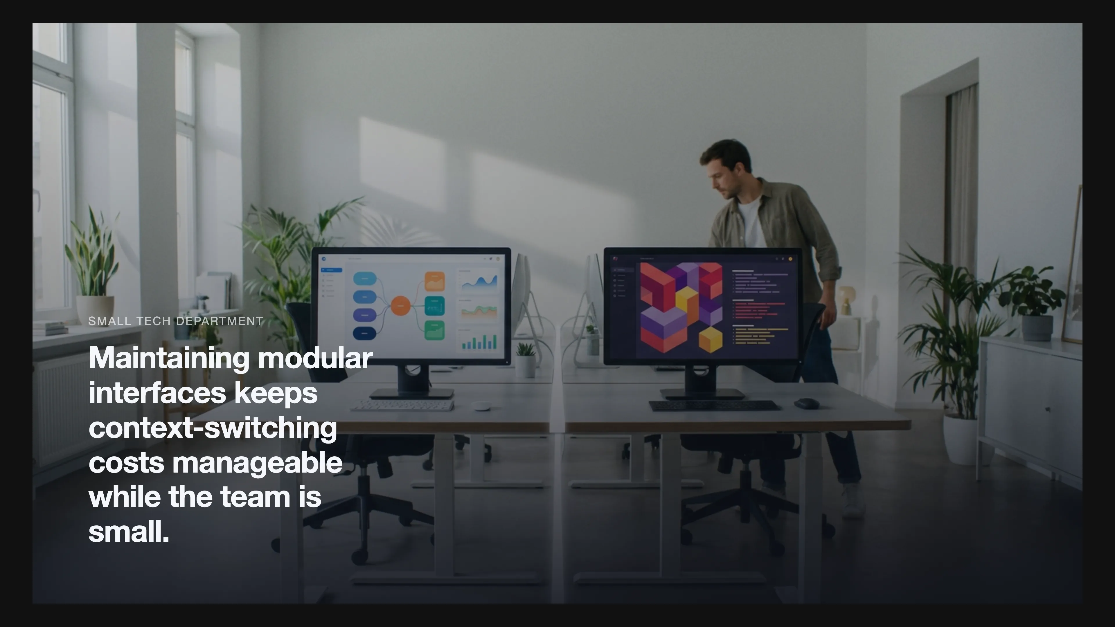 A small tech department workspace with two monitors showing modular interfaces, a clean white line dividing the desk, and an engineer moving between tasks.